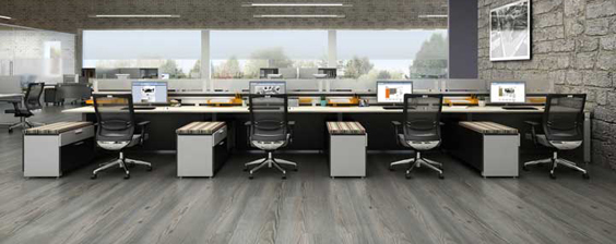 A collaborative workstation featuring a long, low-profile desk with a woodgrain laminate surface and white metal legs. The desk is flanked by two sets of open shelving, providing ample space for storage and organization.